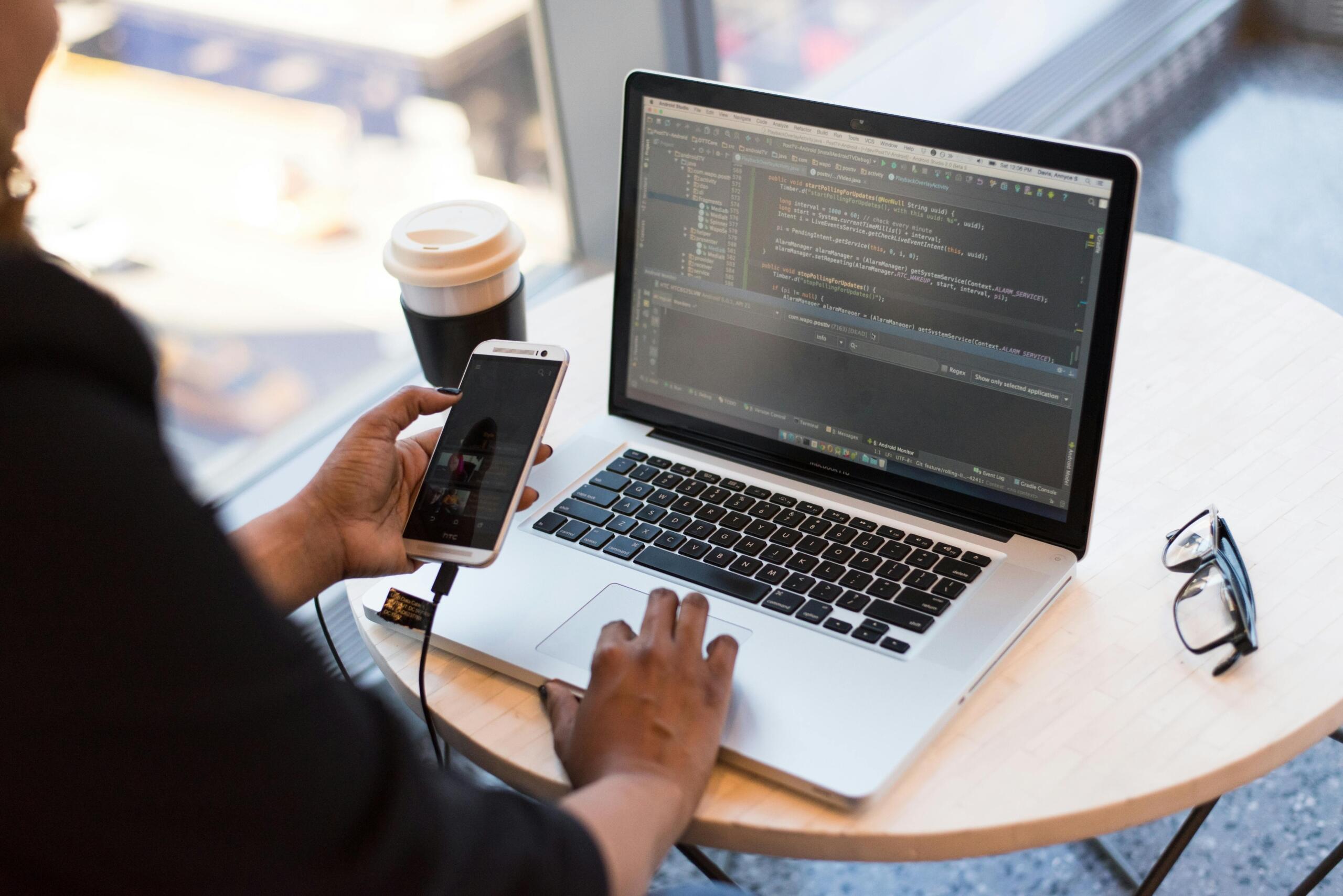 Person holding a smartphone while working on a laptop displaying programming code.