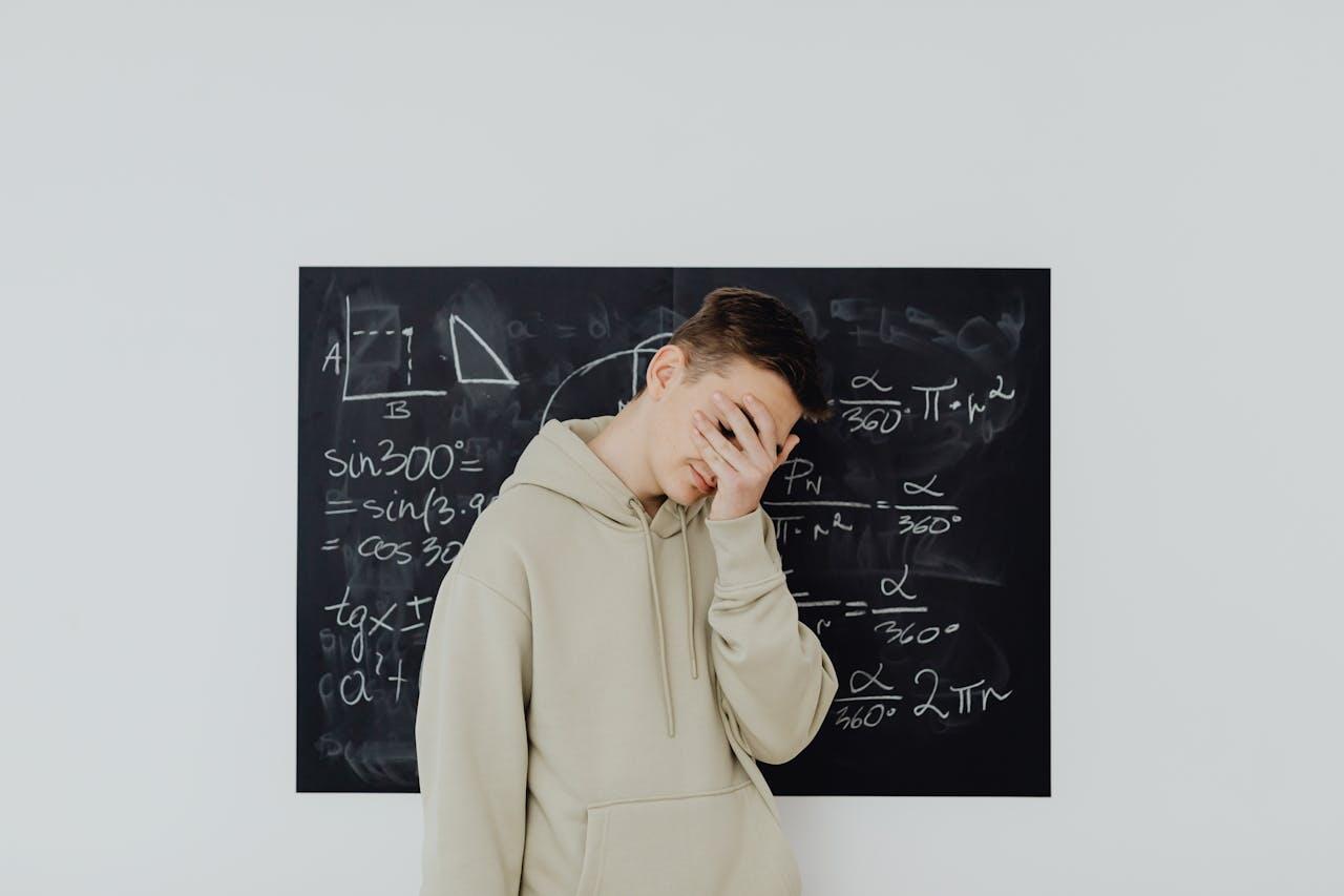 Boy in beige hoodie covering his face in front of a blackboard filled with math notes 