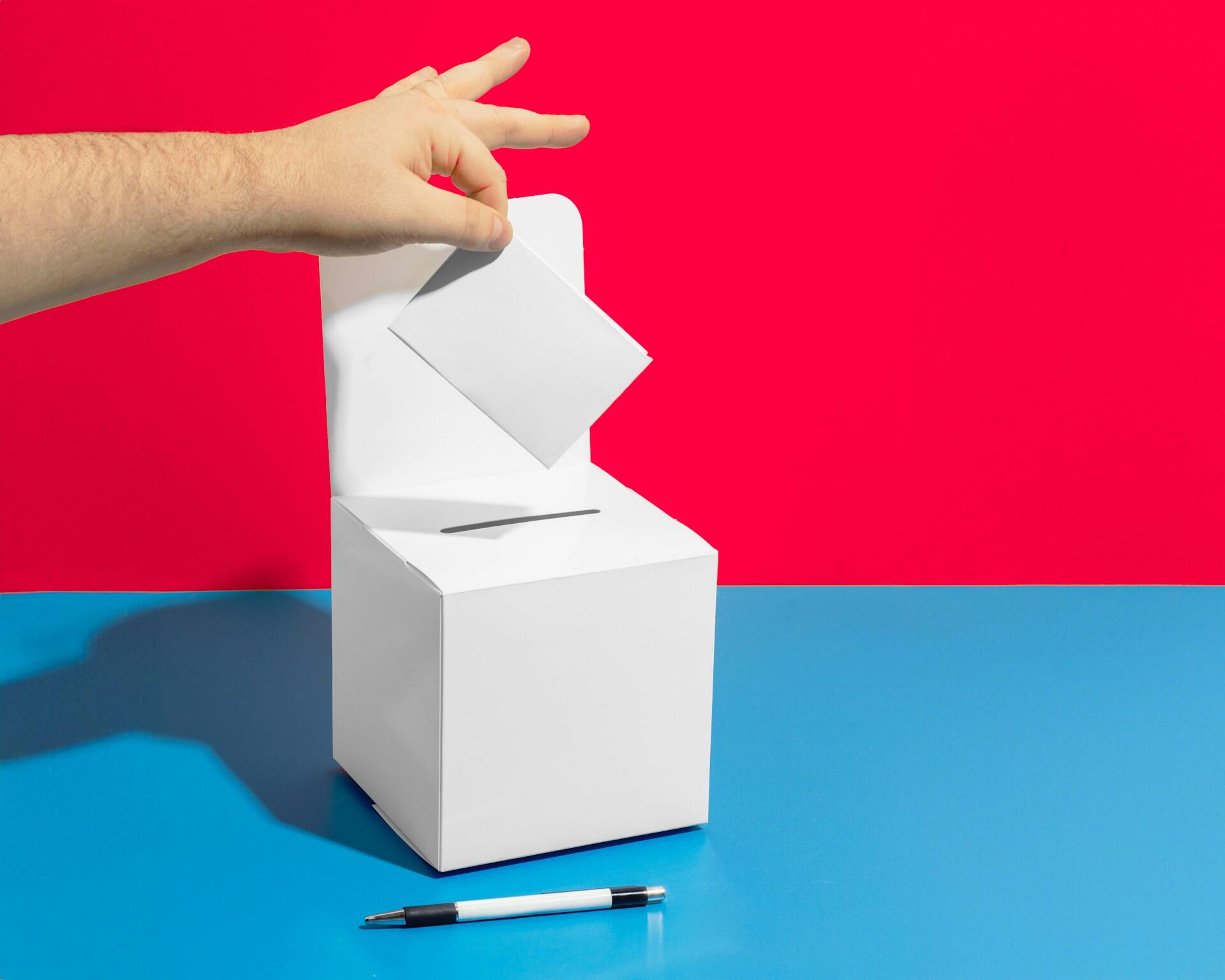 A hand holds a paper over a ballot box.