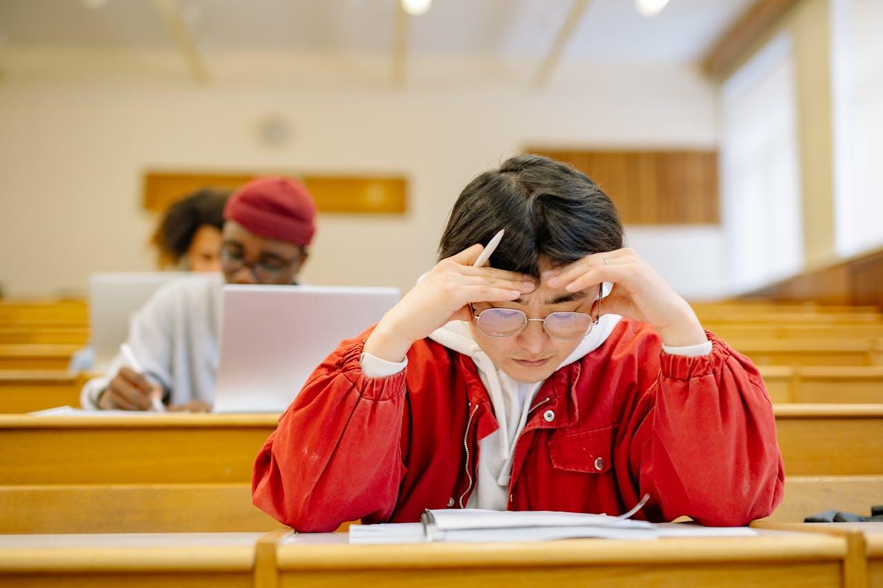 A student in a red jacket rests their head on their hands, appearing stressed, with classmates and laptops in a lecture hall.