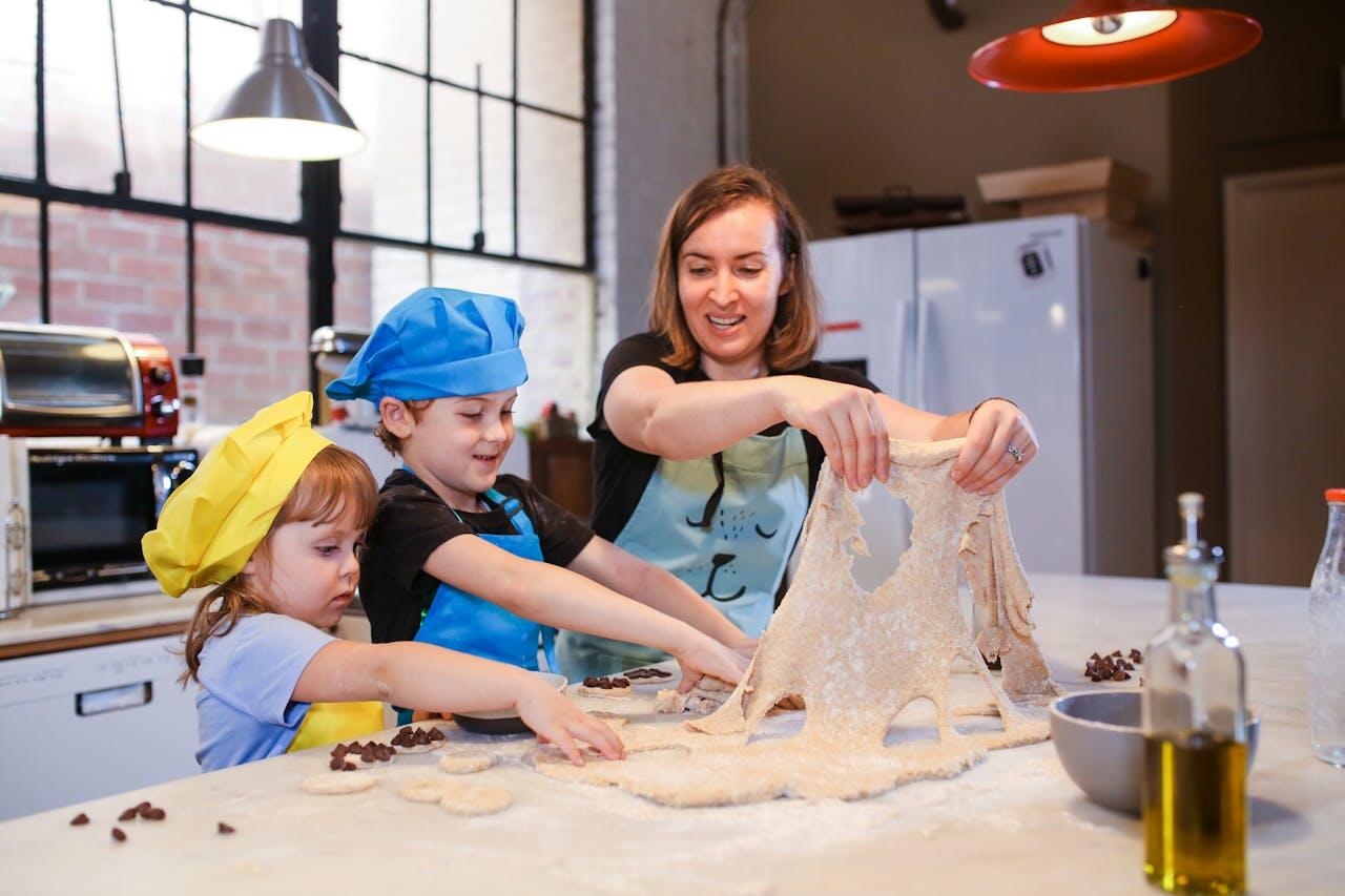 A woman and two children in colorful aprons and hats playfully stretch dough on a kitchen counter, surrounded by baking ingredients.