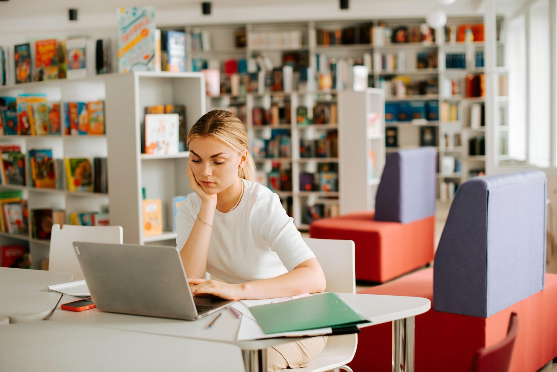 Person using a laptop computer in a library with bookshelves and colorful furniture.