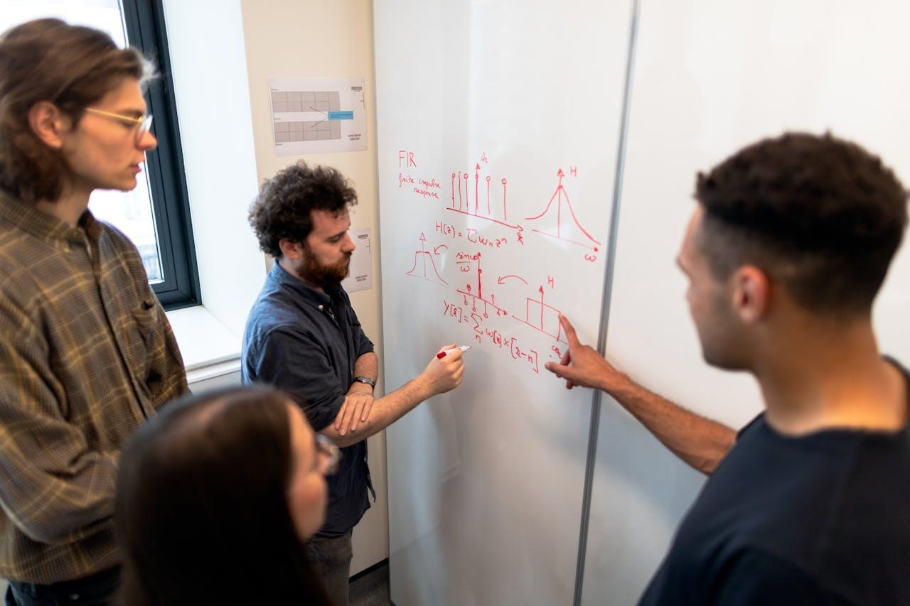 A group of students collaboratively analyze formulas and diagrams on a whiteboard, engaged in a discussion about signal processing concepts.