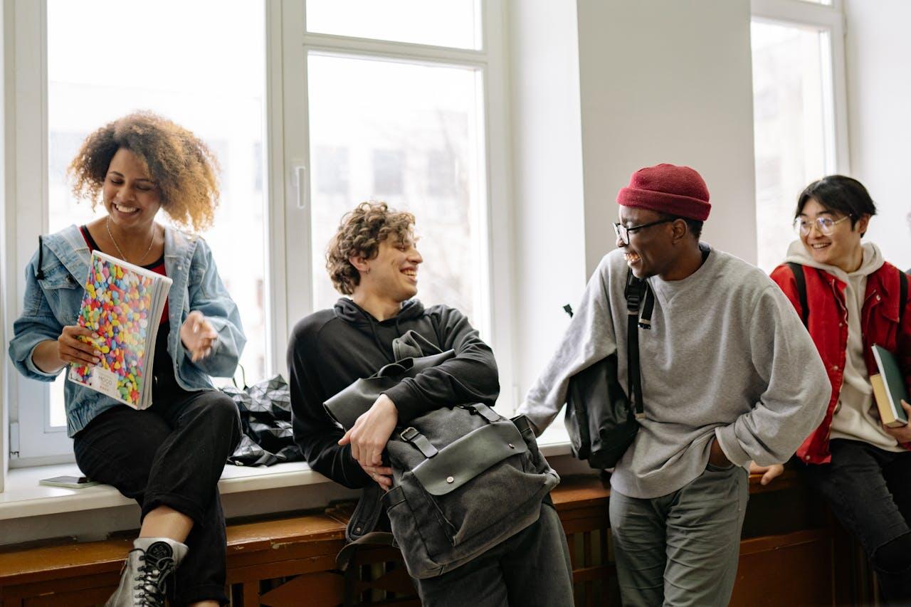 A group of four students engages in conversation by a window, with one holding a colorful notebook and various backpacks visible.