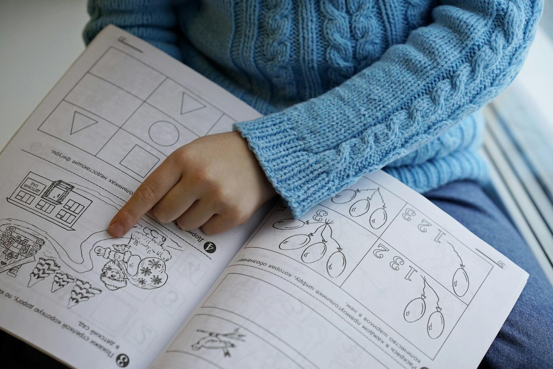 A child's hand points to a coloring page in a workbook featuring a snowy scene with a snowman and decorated trees, while wearing a blue sweater.
