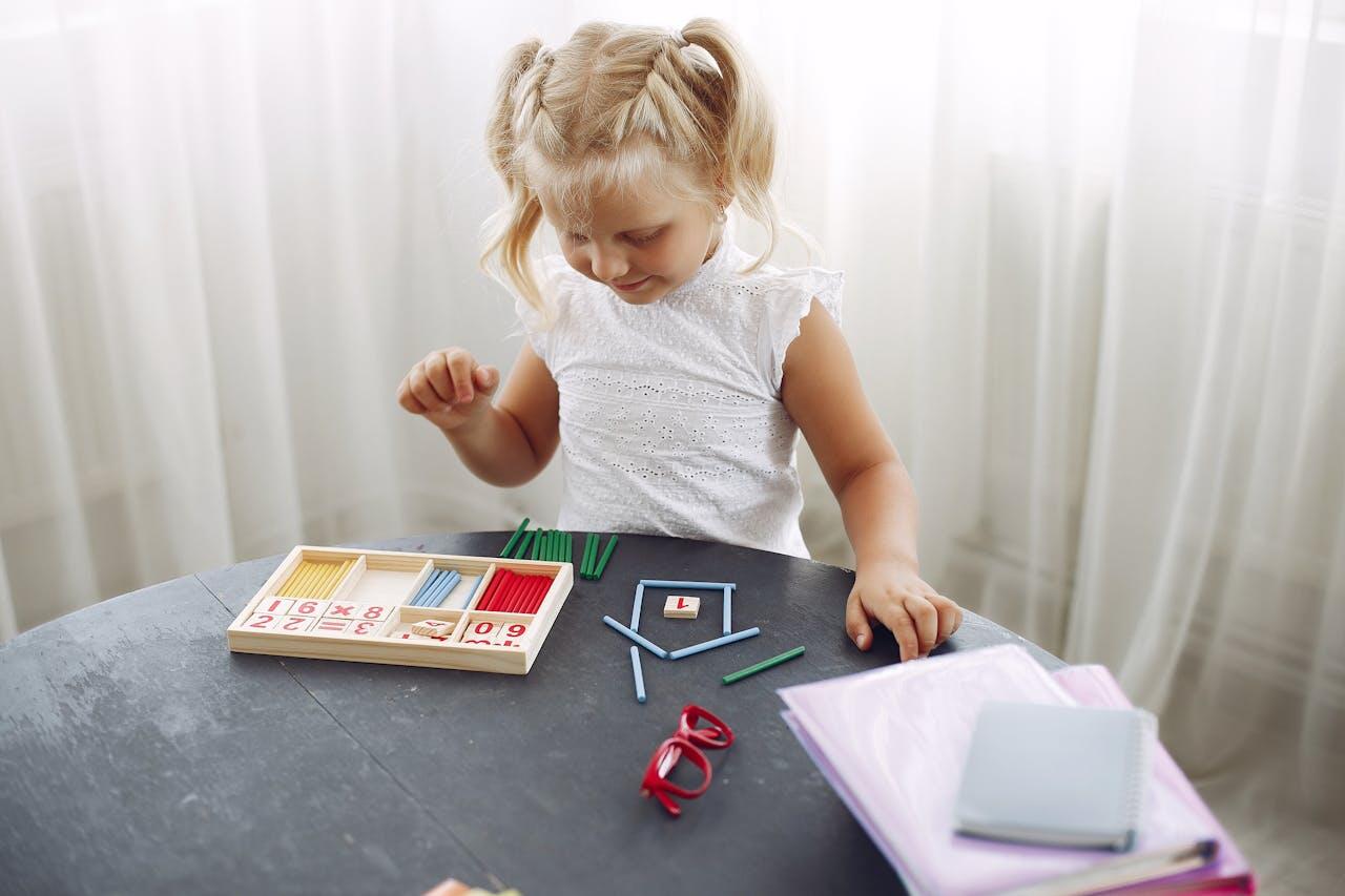A child plays with colorful educational toys on a dark table, surrounded by notebooks and school supplies, in a bright room.
