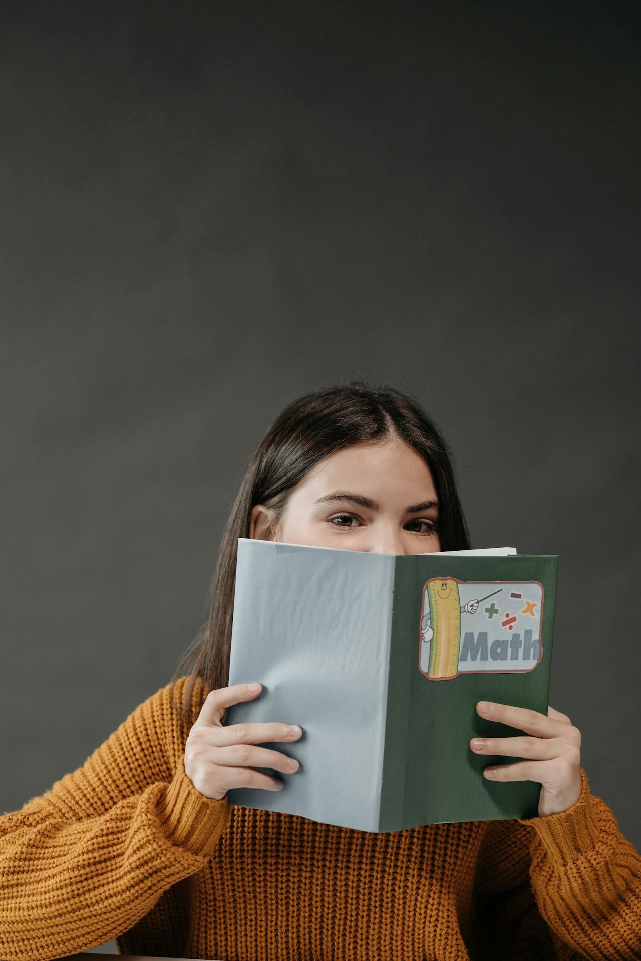 A person in a cozy orange sweater holds a green and blue math book in front of them, with a dark gray background.