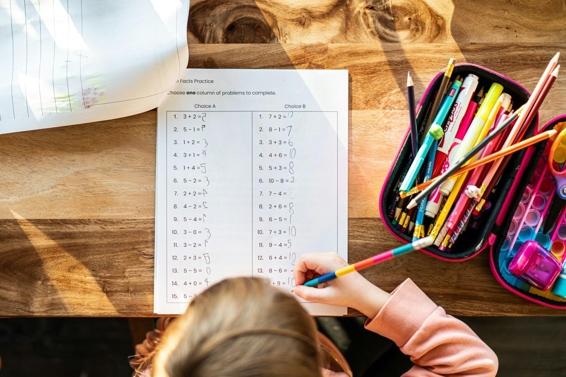 A child works on a math worksheet, using colorful pens, with a pencil case nearby on a wooden table. Natural light illuminates the scene.