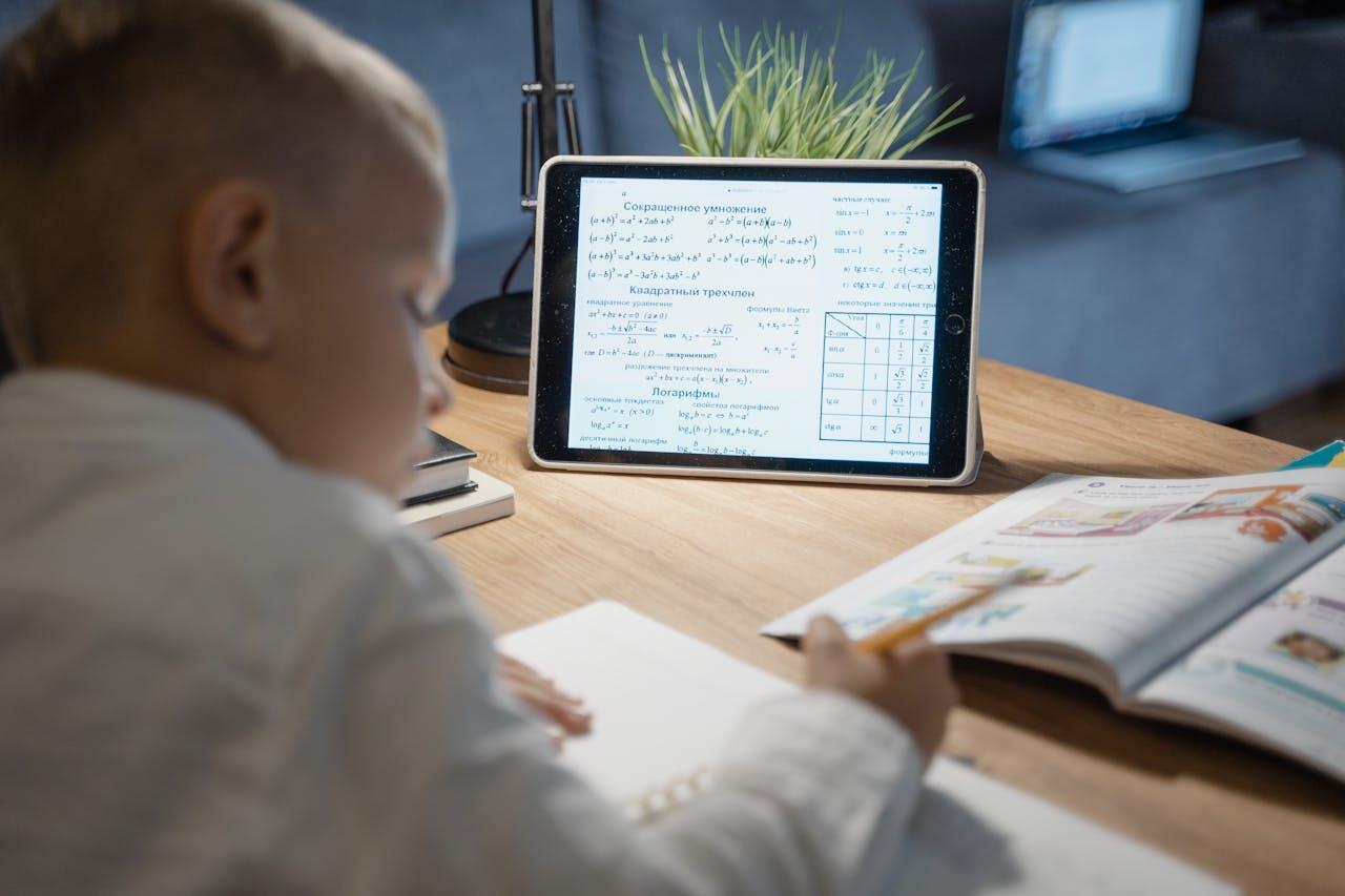 A child studies math on a tablet at a desk, surrounded by notebooks and a plant, with a laptop visible in the background.