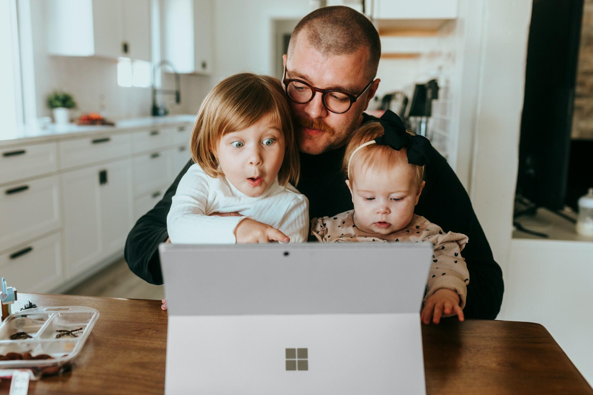 A parent and two children interact with a tablet on a wooden table in a bright, modern kitchen.