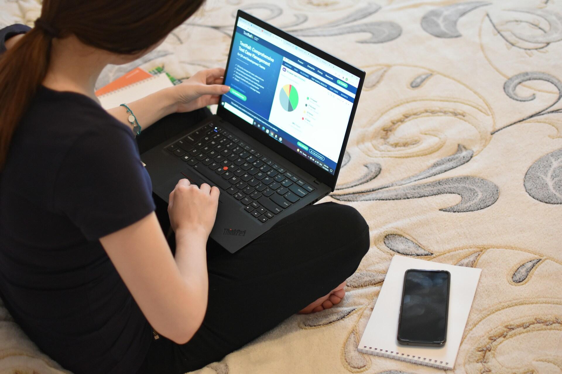 A woman sitting cross-legged on a bed using a laptop, viewing data on a website, with a notebook and smartphone nearby.