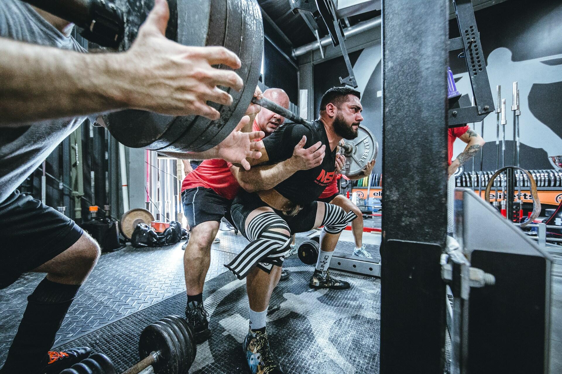 A weightlifter performs a squat with a heavy barbell, supported by two trainers in a gym setting.