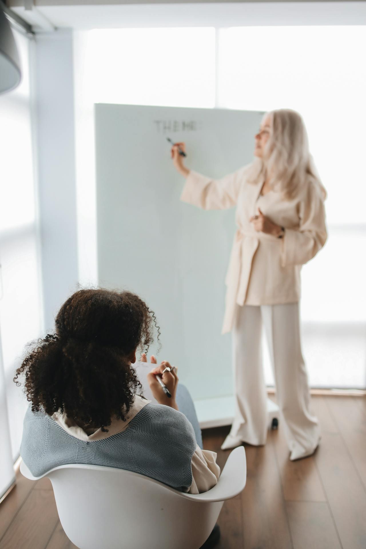 A woman in a cream outfit writes on a whiteboard while another person, seated, takes notes in a modern office space.