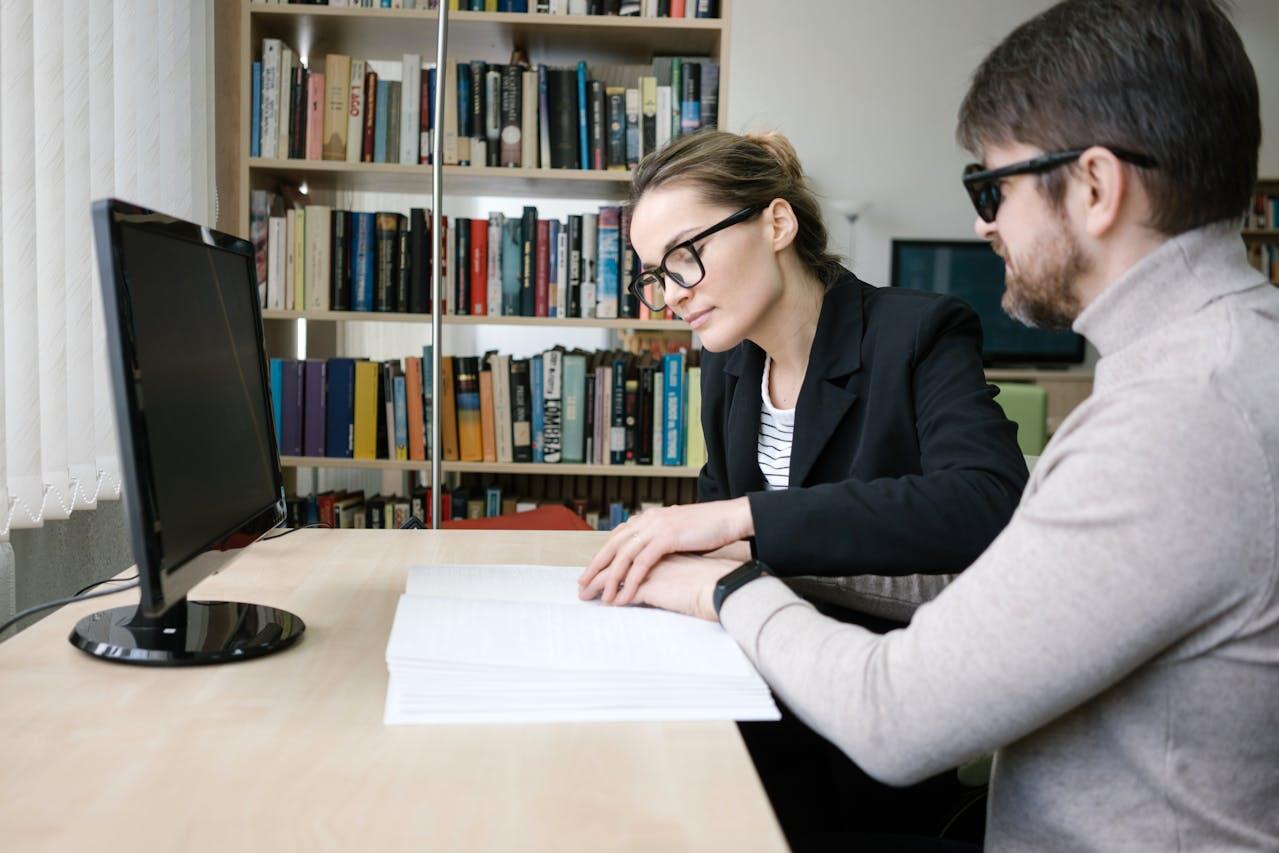 Two individuals are seated at a desk, engaged in discussion over a stack of papers, with a computer monitor visible nearby and shelves of books behind.
