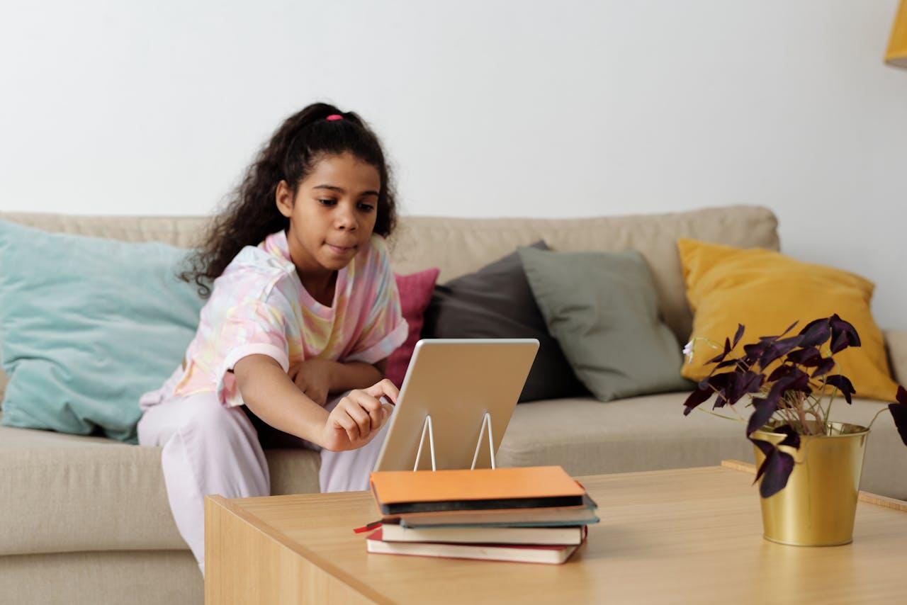 A child sits on a couch, reaching toward a tablet on a coffee table surrounded by books and a potted plant. Colorful cushions are in the background.