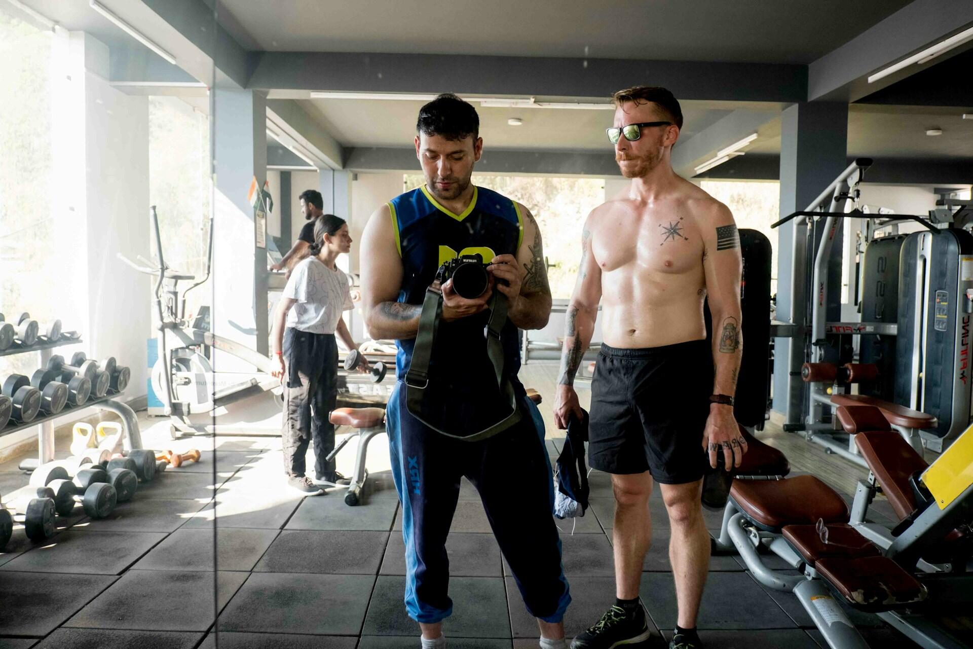 Two men pose in a gym mirror; one holds a camera while the other stands shirtless with tattoos, surrounded by gym equipment.