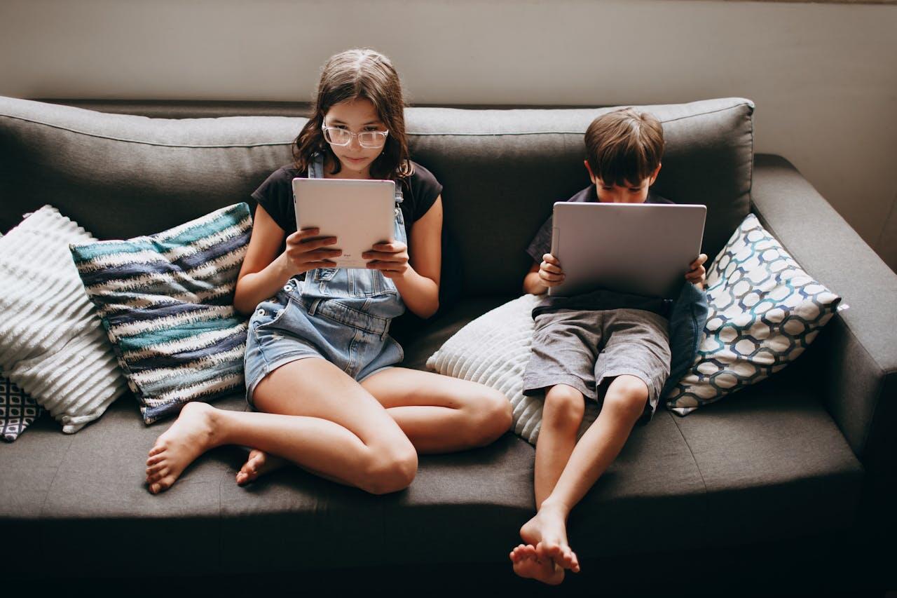 Two children sit on a couch, each absorbed in their tablets. Surrounding them are colorful cushions in a cozy indoor setting.