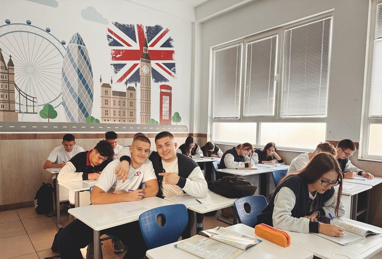 Students engaged in writing activities in a well-lit classroom adorned with a mural of London landmarks and a British flag.