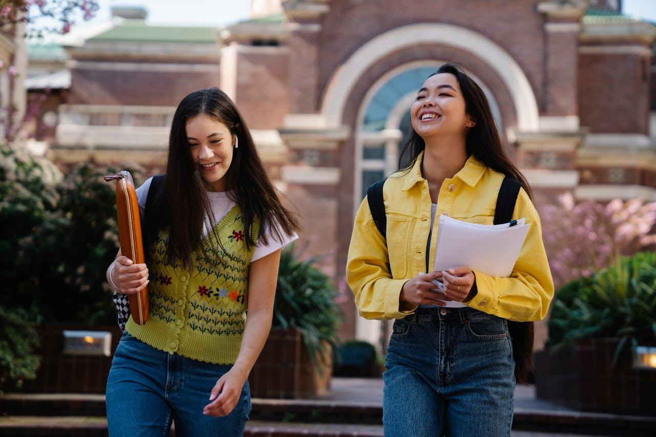 Two young women walk side by side on a campus, one holding a notebook and the other with sheets of paper, surrounded by greenery and historic architecture.