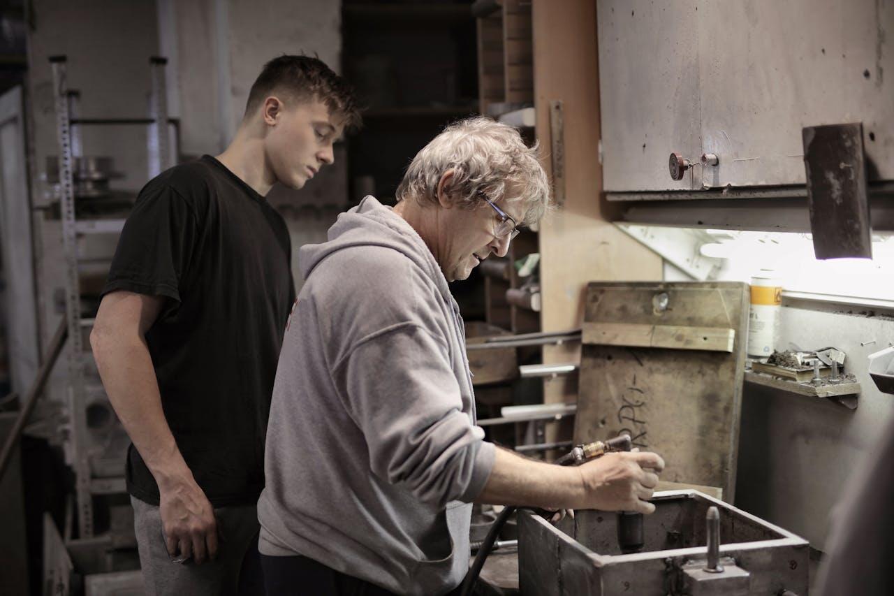 A man in a gray sweatshirt works with tools in a cluttered workshop while another person observes closely.