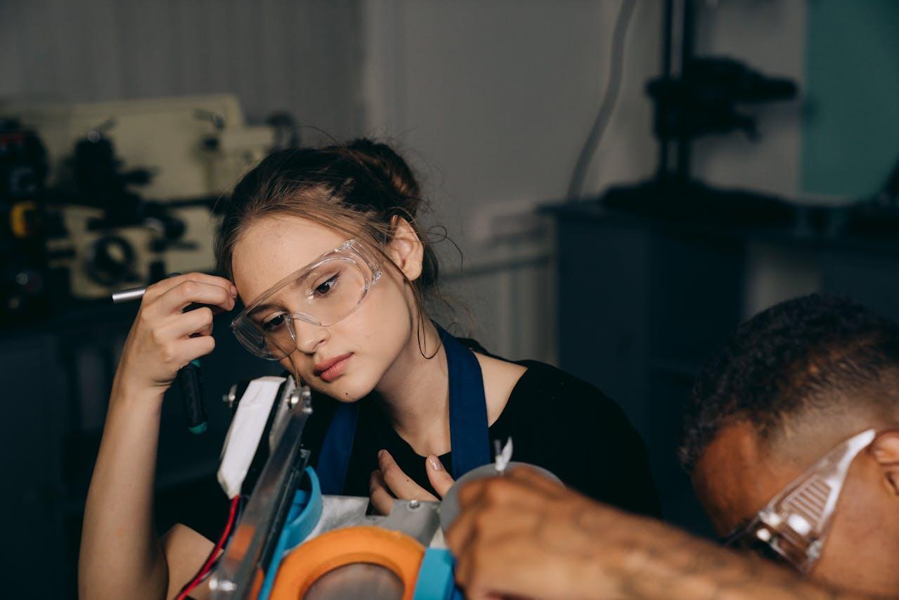 A person wearing a blue apron works intently with tools on a machine, surrounded by various equipment in a workshop setting.