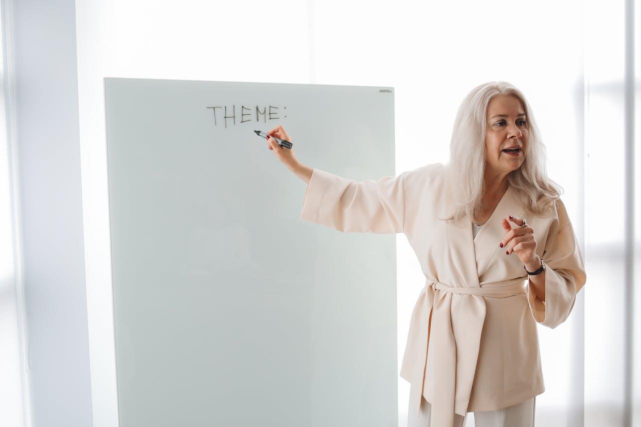 A woman in a light beige outfit writes "THEME:" on a whiteboard, gesturing as she leads a discussion in a bright room.