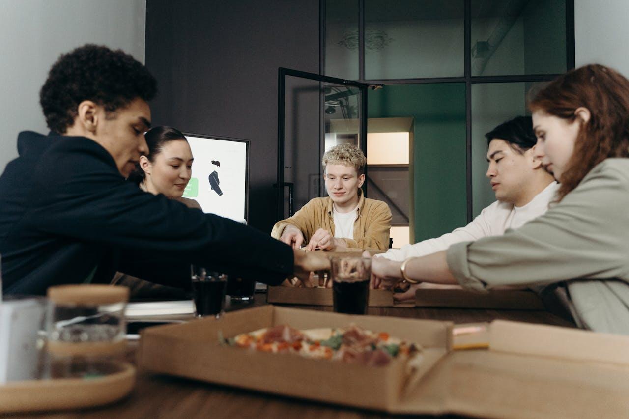 A group of people engaged in a collaborative meeting, sharing ideas over pizza boxes and drinks in a modern office setting.