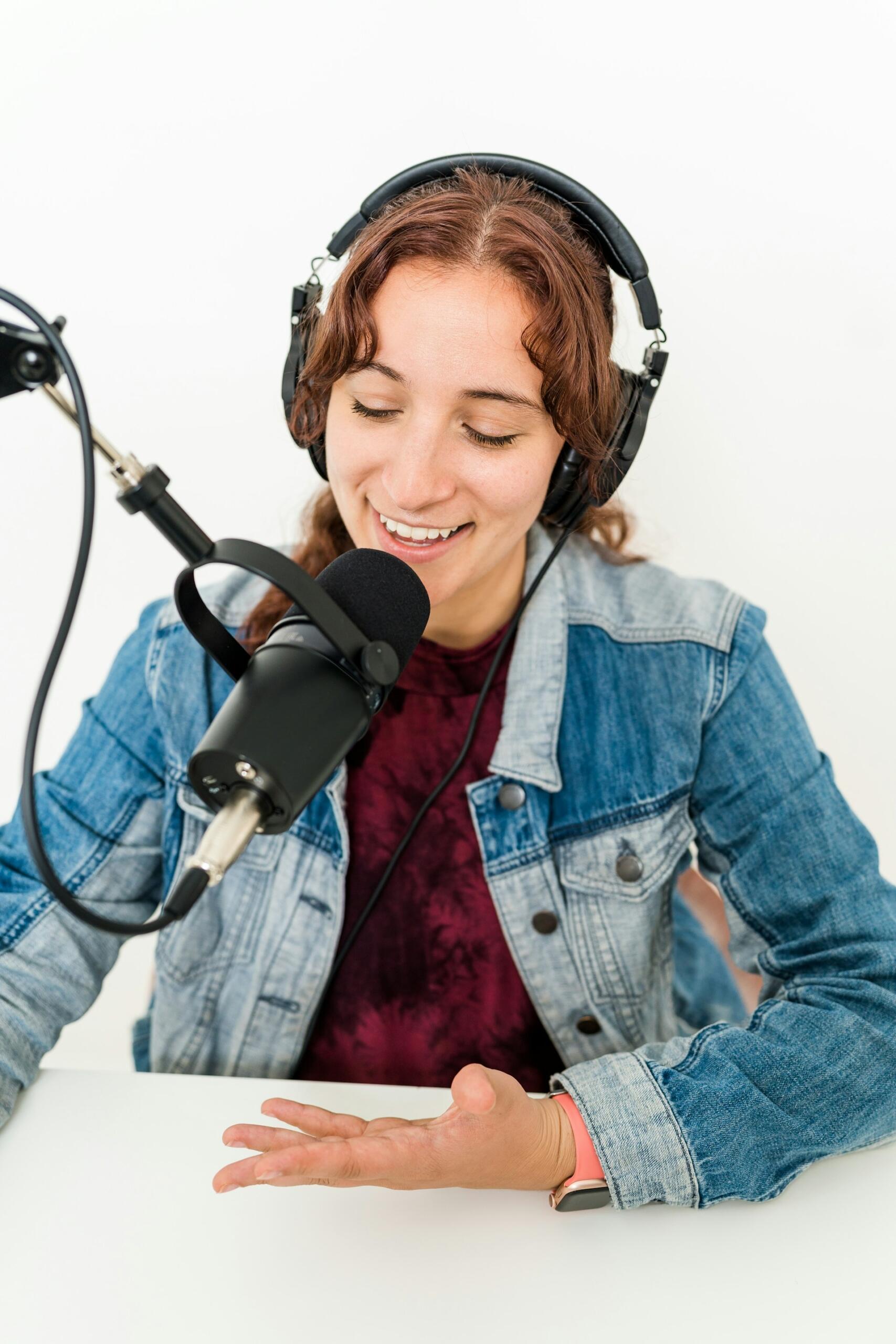 A person wearing headphones sits at a table, gesturing with their hand towards a microphone, dressed in a denim jacket over a dark shirt.