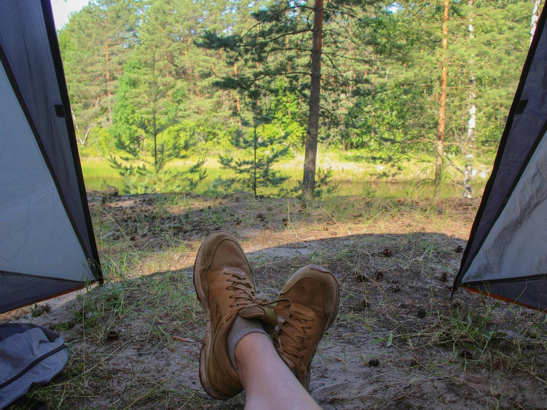 A person's feet in rugged boots resting outside a tent, with a lush forest and calm water visible in the background.