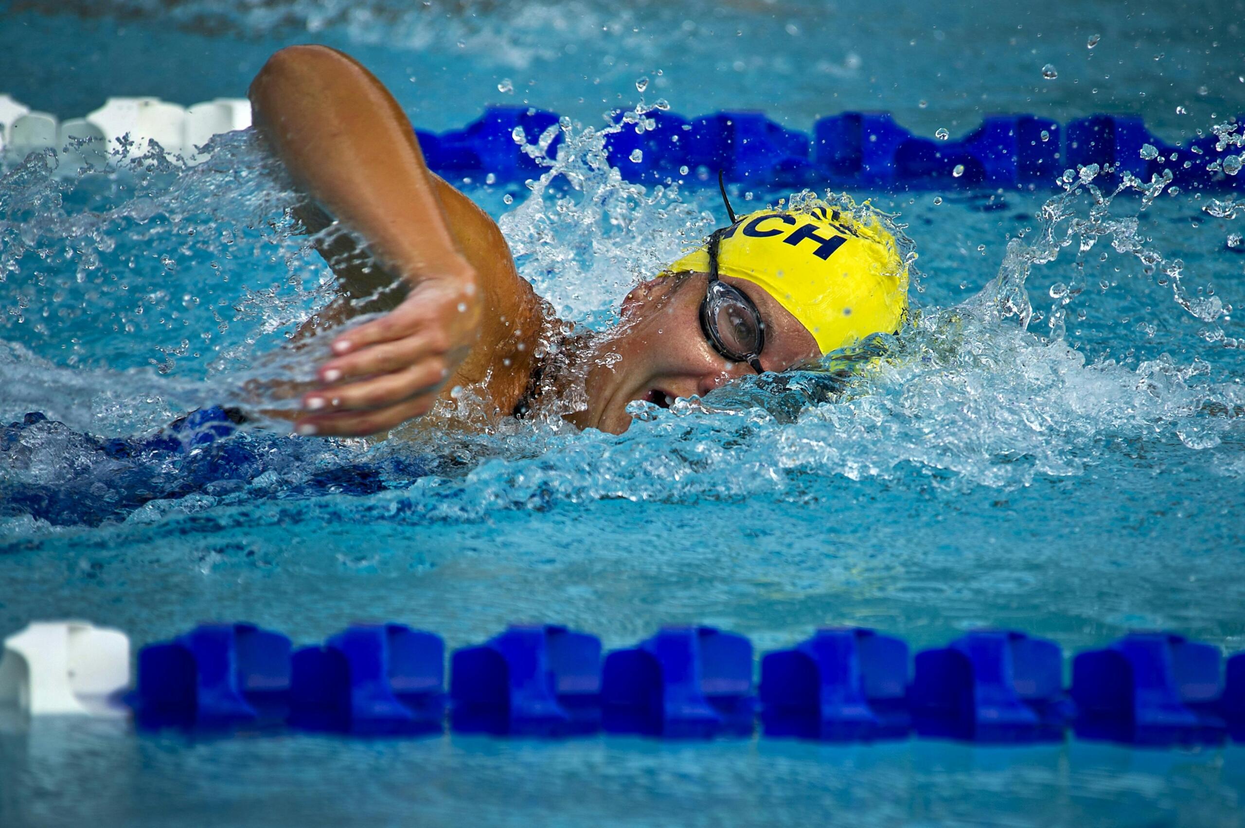 A swimmer wearing a yellow cap floats near the edge of a clear blue swimming pool.