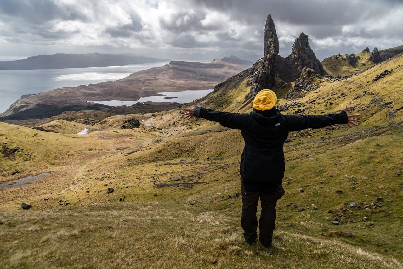 A person in a yellow hat stands with arms outstretched, overlooking rugged hills and a serene coastline under a cloudy sky.