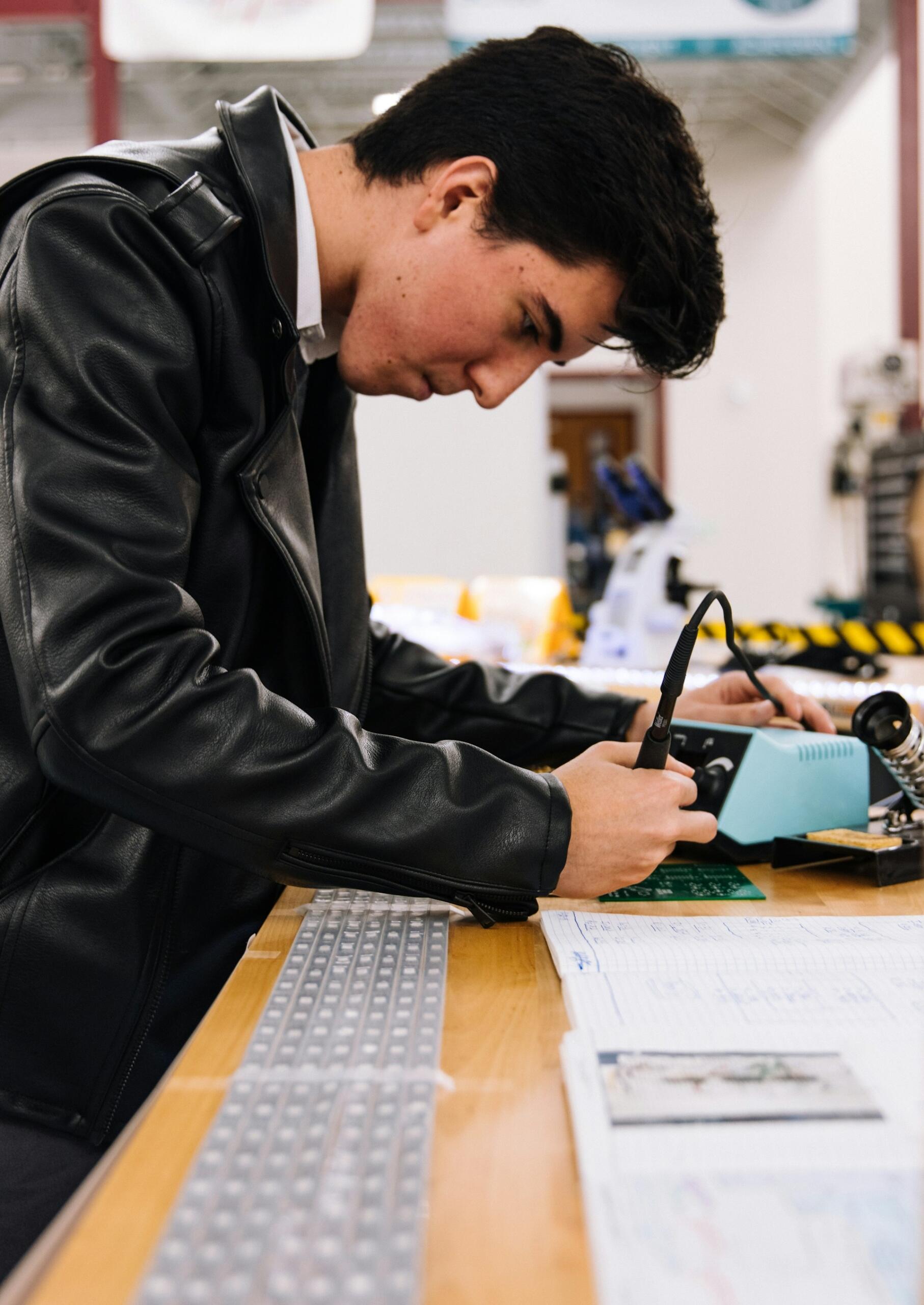 A person in a black leather jacket works intently at a wooden table with tools and printed circuit boards. Papers are scattered nearby.