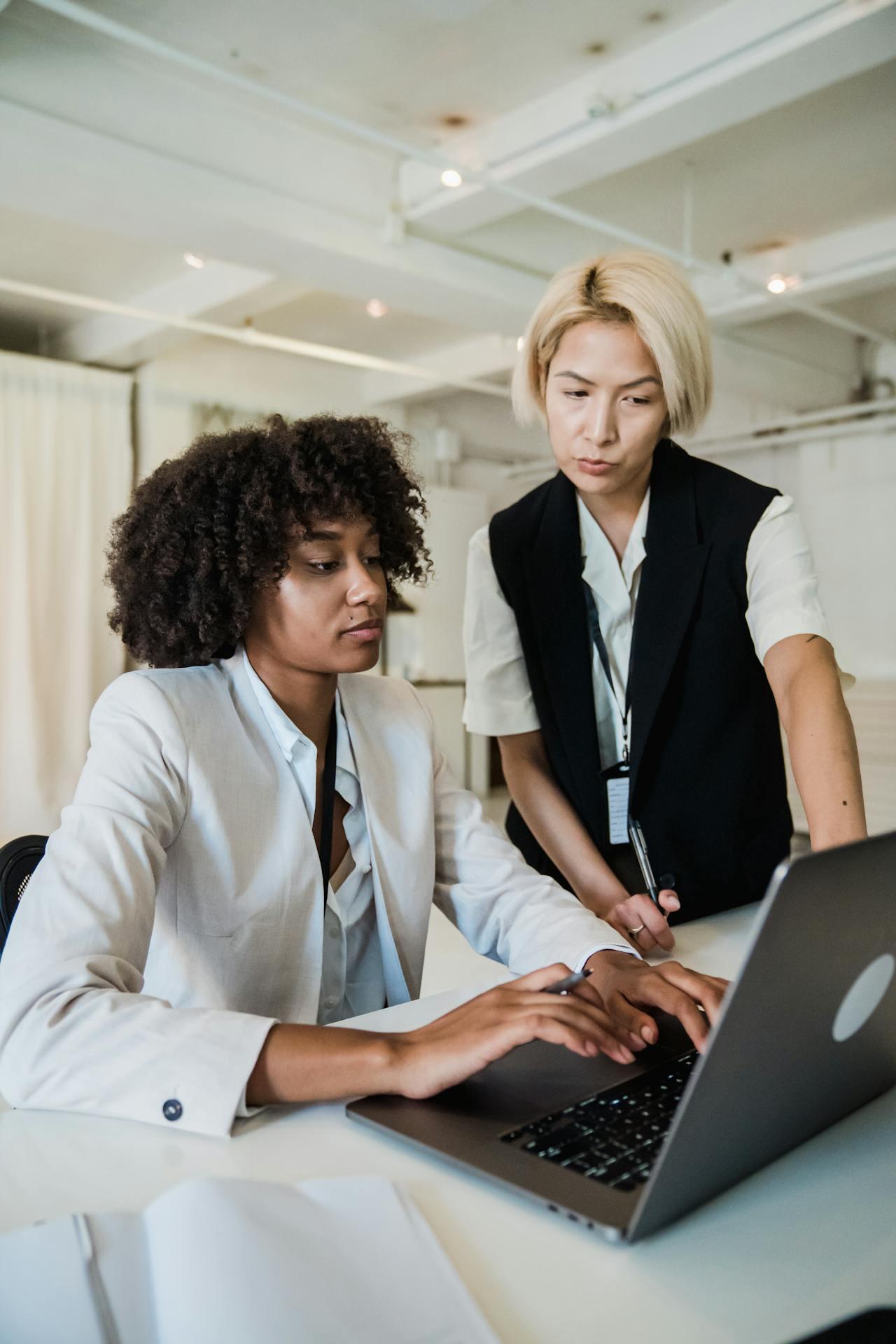 Two women collaborate at a desk, one focused on a laptop while the other gestures toward the screen, in a modern office setting.