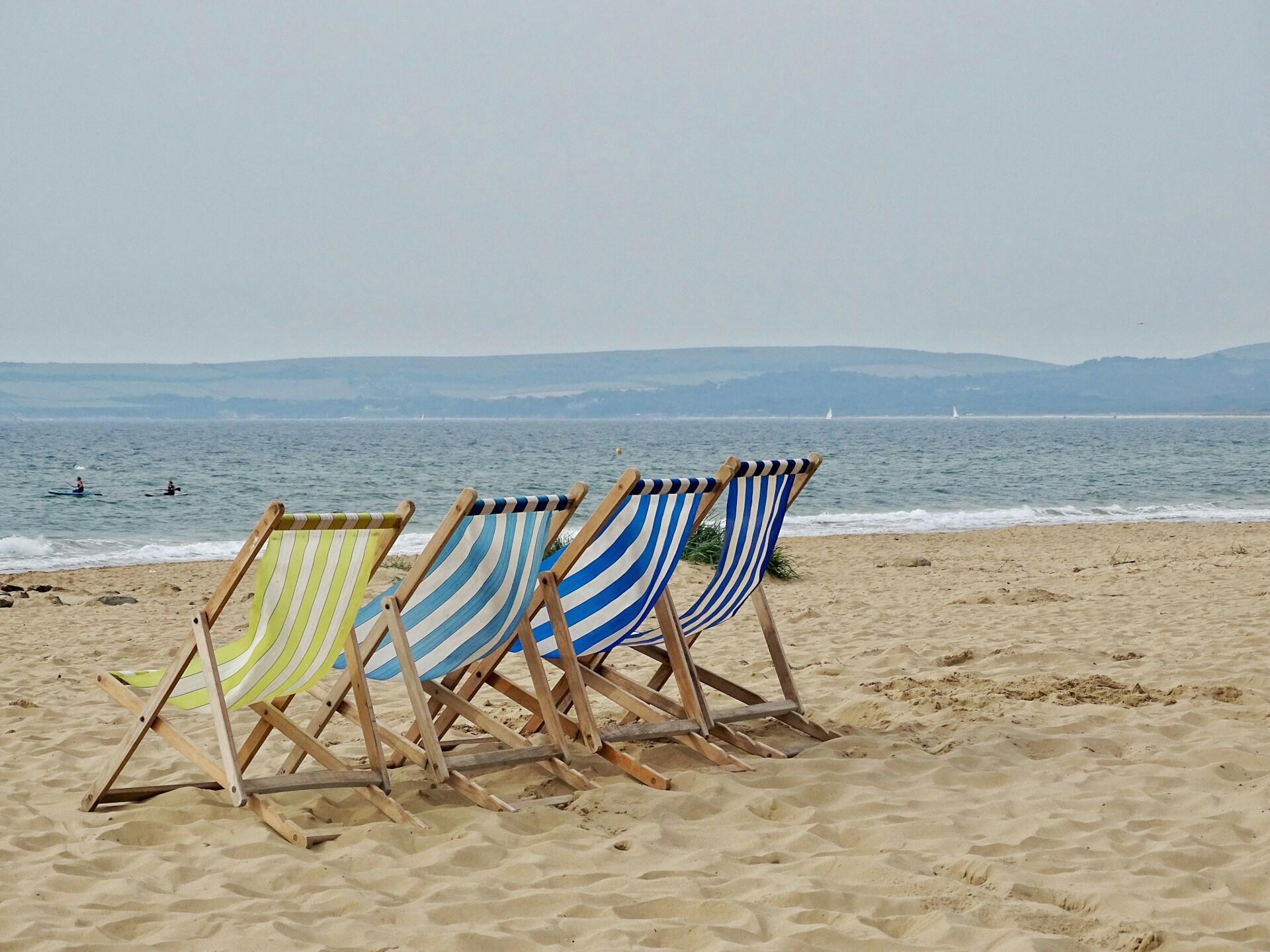 Five striped deck chairs in green, blue, and white face the calm sea on a sandy beach, with kayakers in the distance.