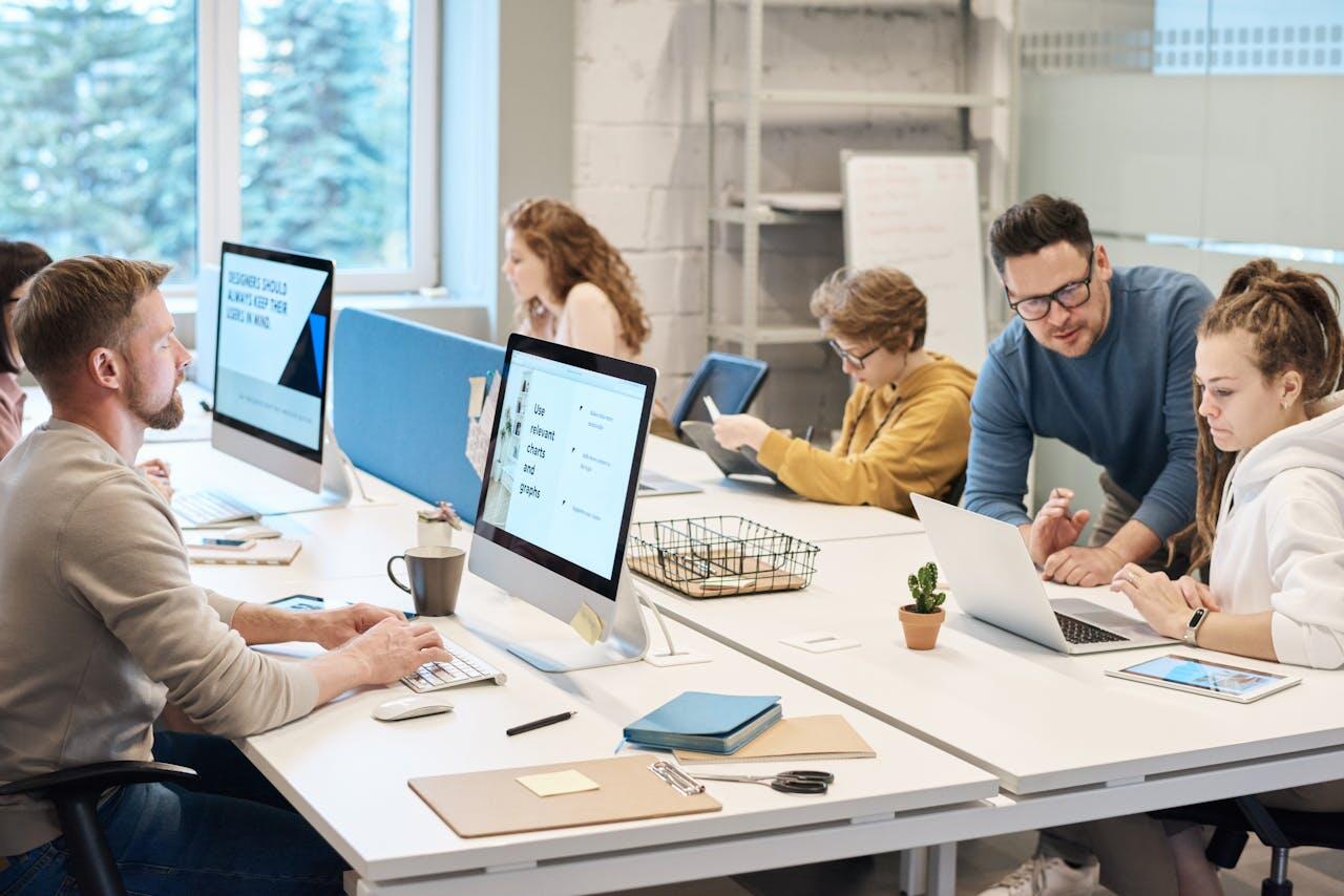 A collaborative workspace with several individuals engaged in discussions and working on computers, surrounded by office supplies and a plant.