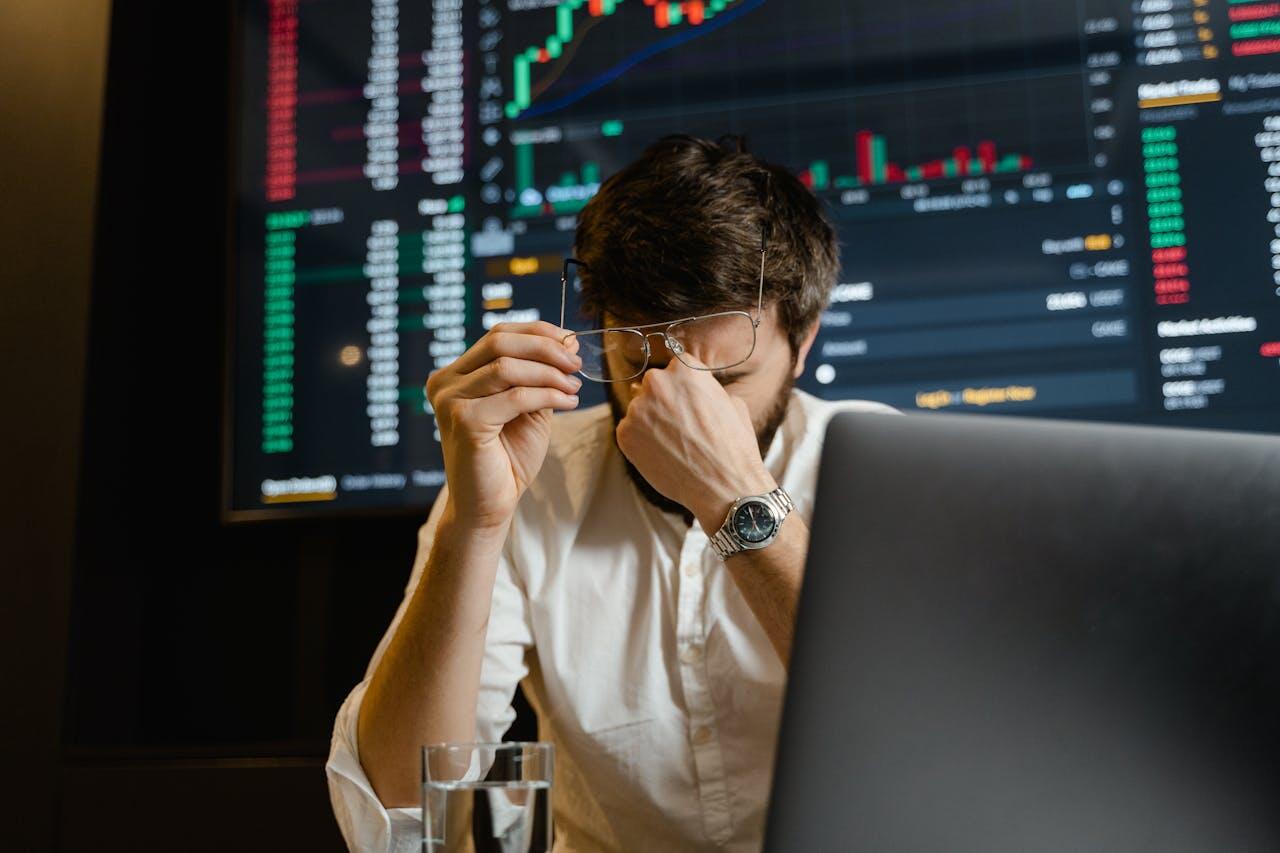 A person in a white shirt sits at a desk with a laptop, analyzing financial data displayed on a large screen.