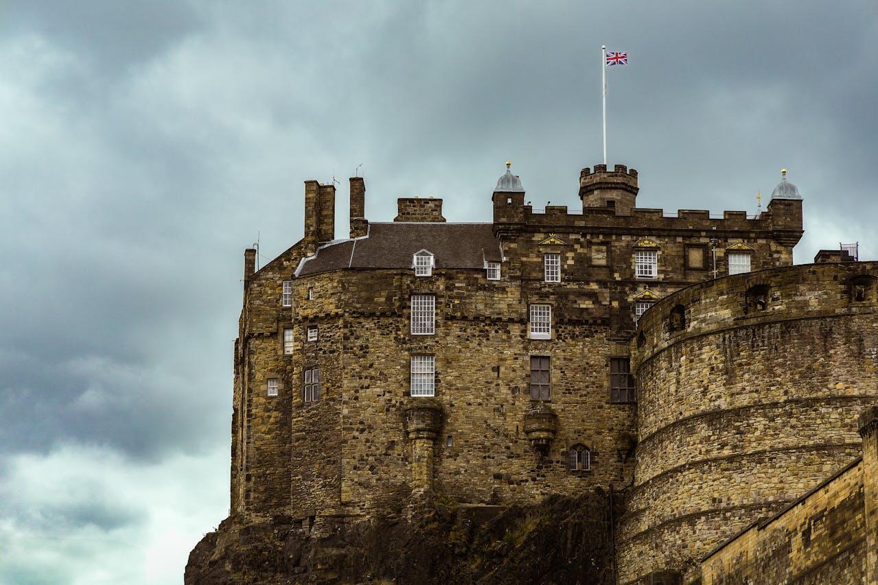 Edinburgh Castle stands majestically on a rocky outcrop, with a British flag fluttering against a cloudy sky.