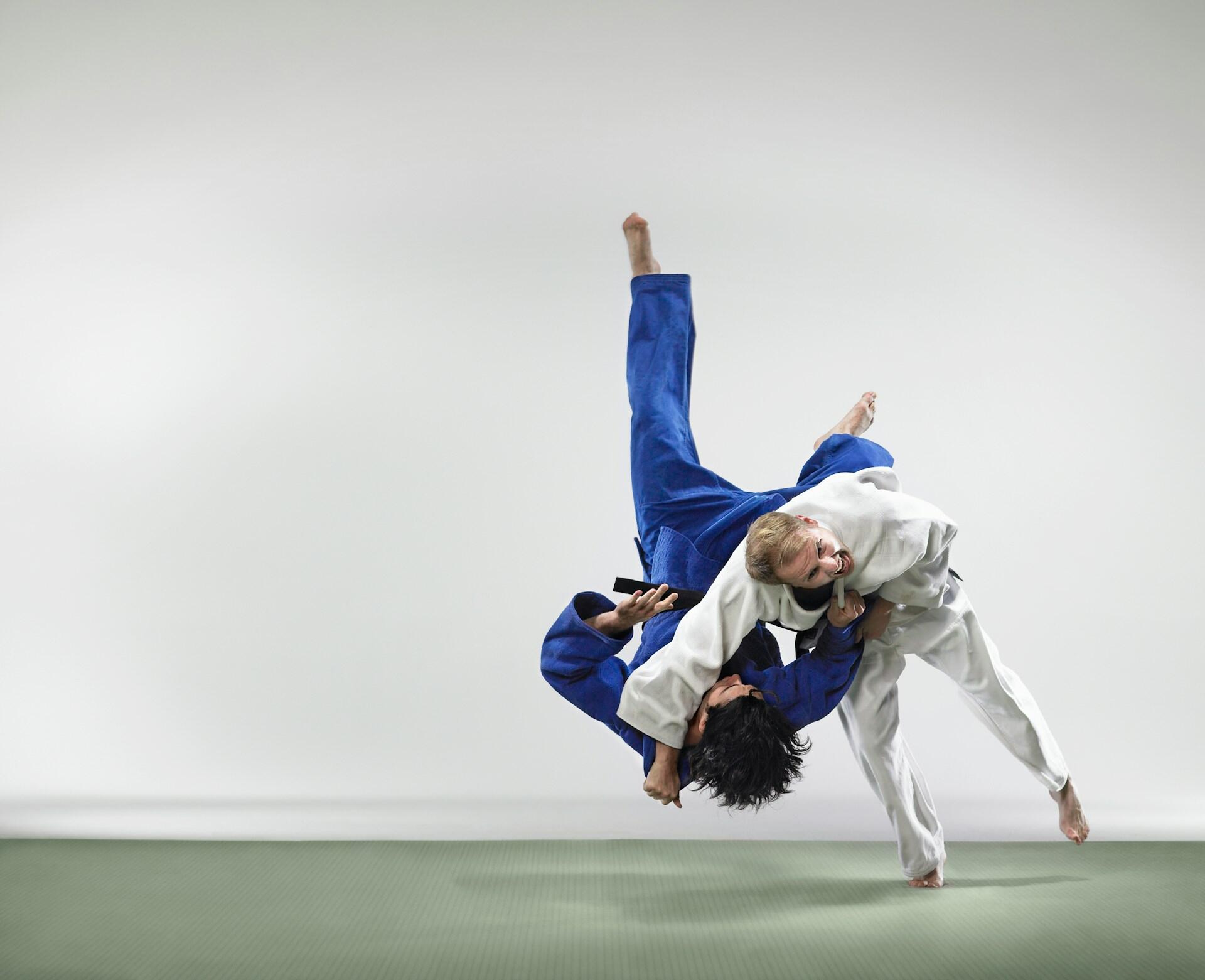 Two judo practitioners in blue and white outfits executing a split on a training mat in a dojo setting. 
