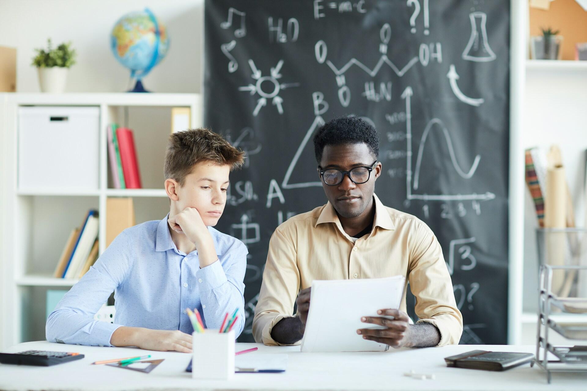 A person in a classroom considers a paper while a student looks on. 