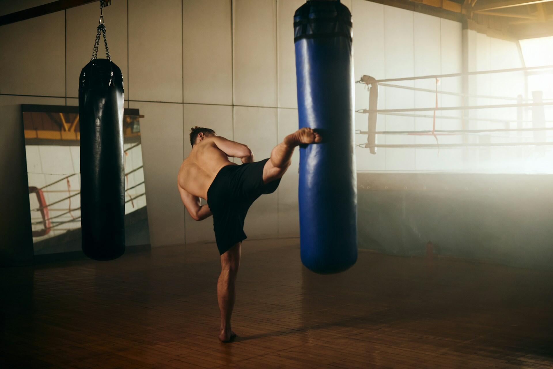 A man delivering a powerful kick to a punching bag in a well-equipped gym setting.