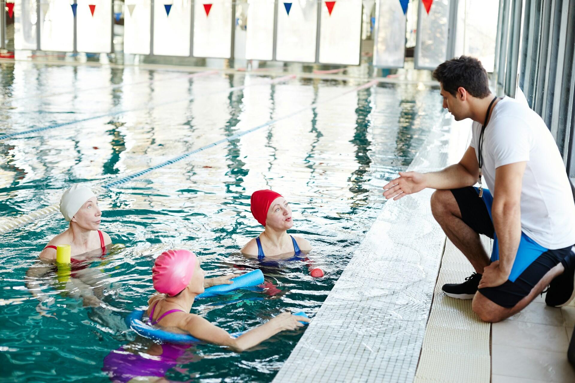 three elderly women swimming in a pool discussing with a lifeguard
