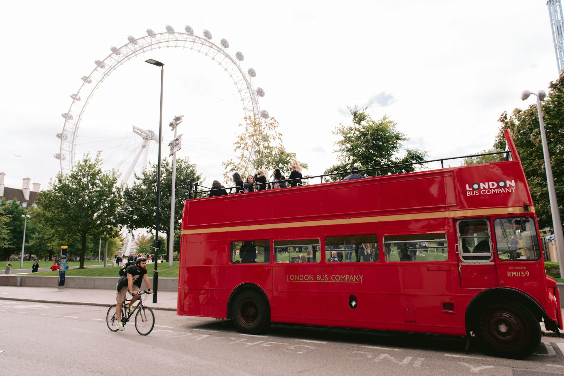 A classic red double-decker bus is parked near the London Eye, with cyclists and pedestrians enjoying the lively park atmosphere.