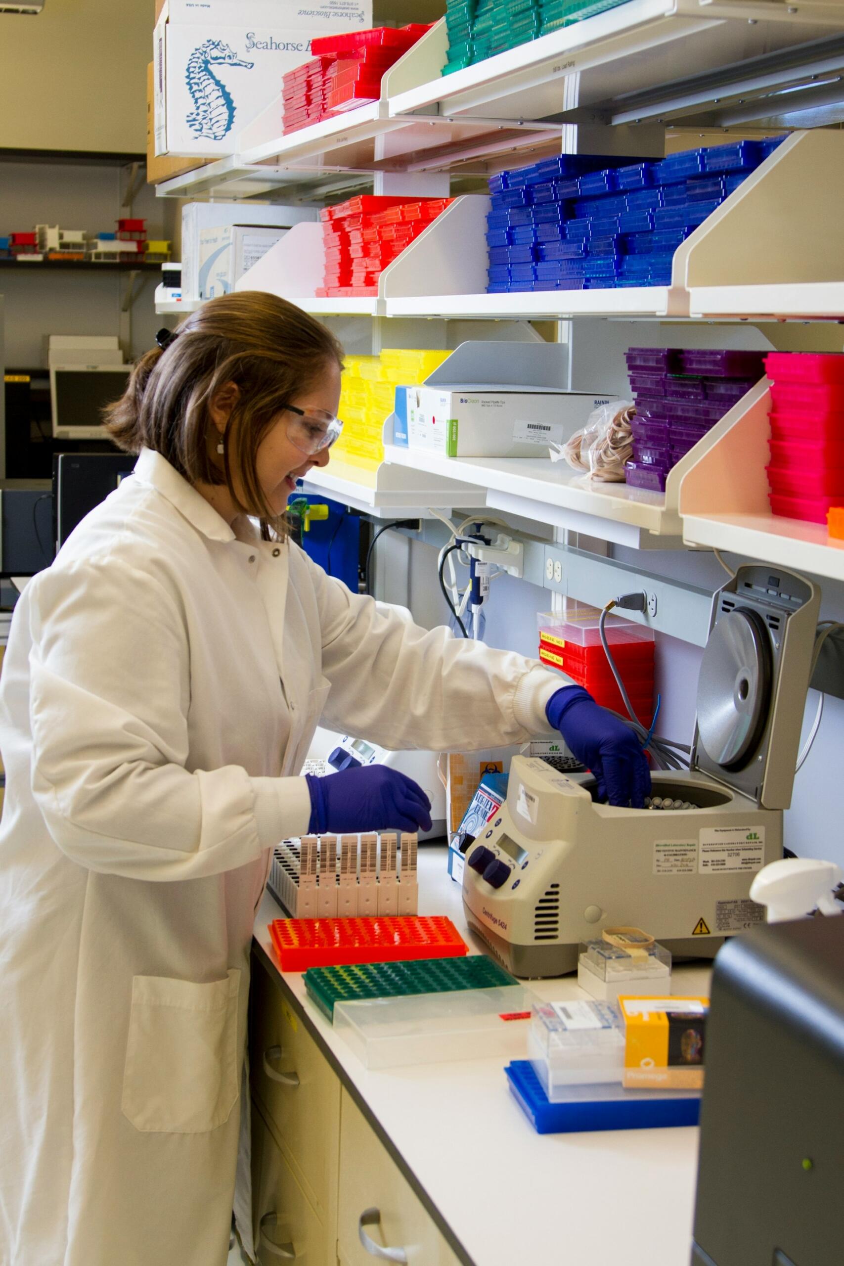 A scientist in a lab coat operates a centrifuge, surrounded by colorful storage boxes and laboratory equipment on a workbench.