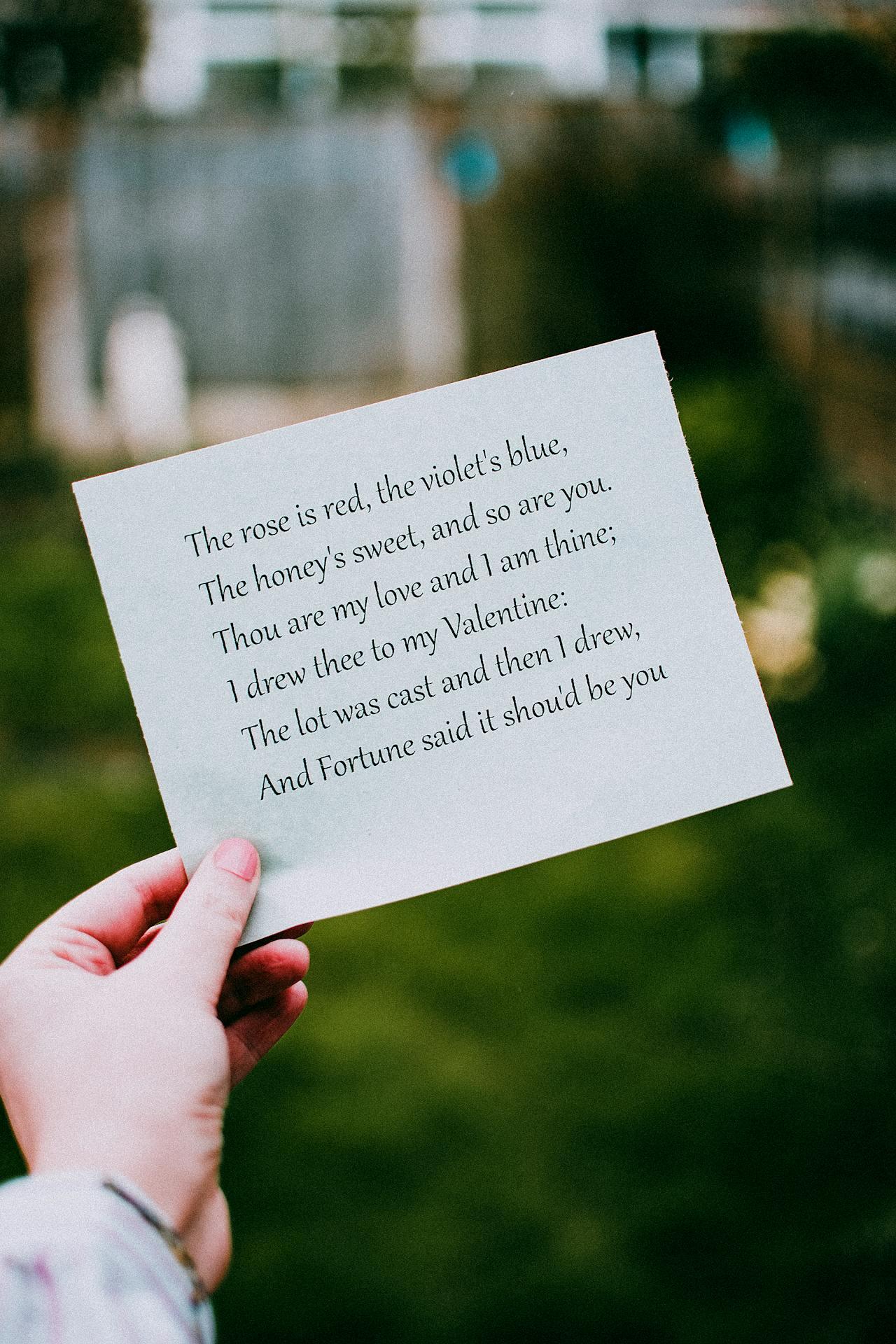 A person holds a paper displaying a romantic poem, with blurred greenery and a fence in the background.