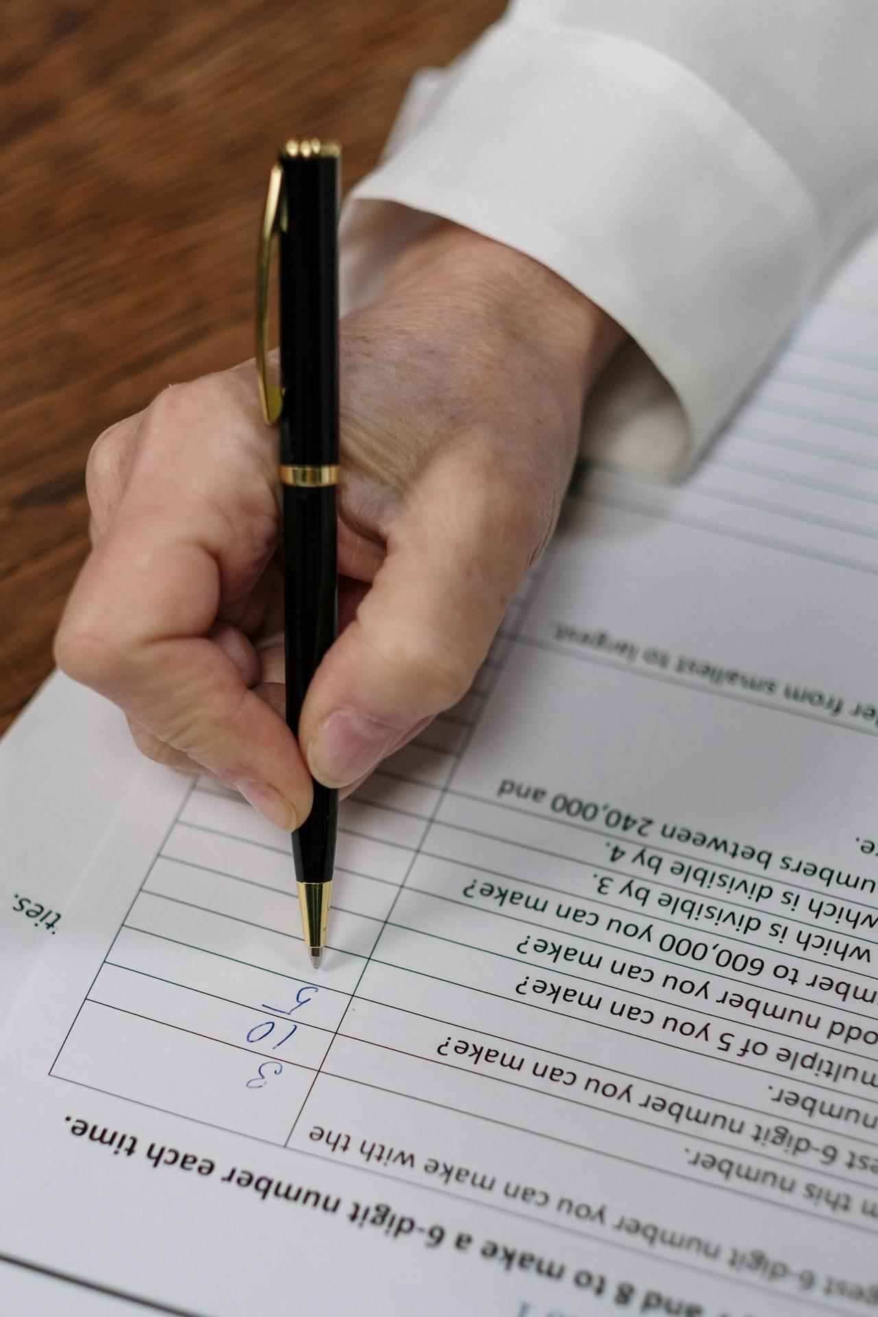 A hand holding a black pen writes on a paper with lists and calculations, focused on numbers in blue ink.