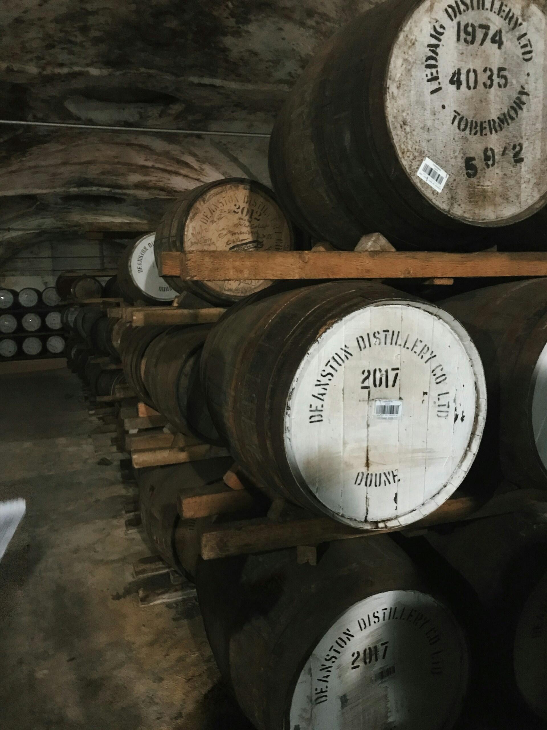 Wooden whiskey barrels stacked in a dimly lit distillery cellar, labeled with dates and the Deanston Distillery logo.
