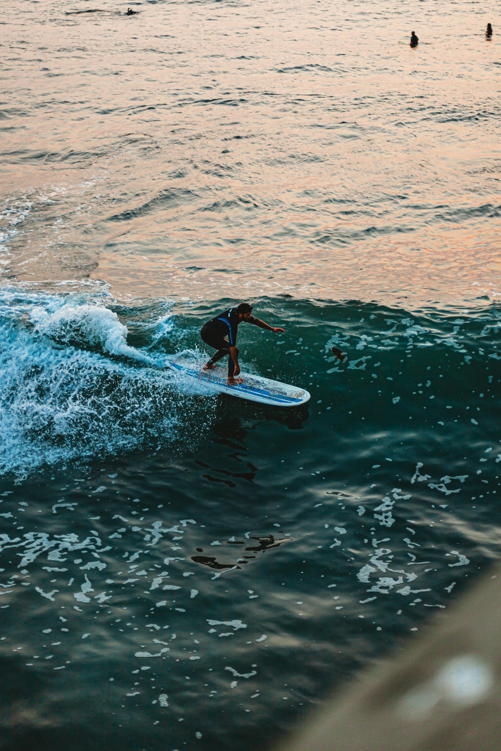 A surfer skillfully rides a vibrant wave at sunset, with calm waters and fellow surfers visible in the background.