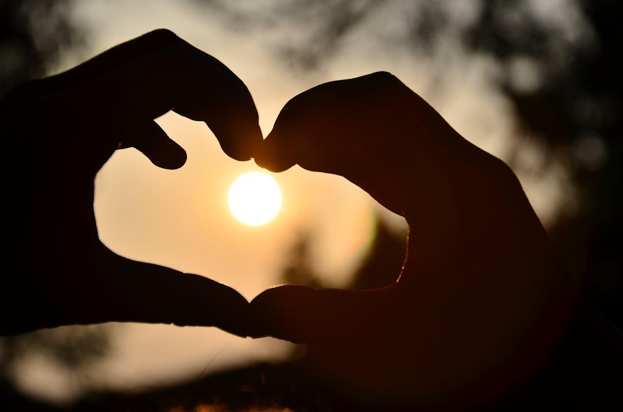Silhouette of two hands forming a heart shape against a sunset, with the sun glowing brightly in the center.
