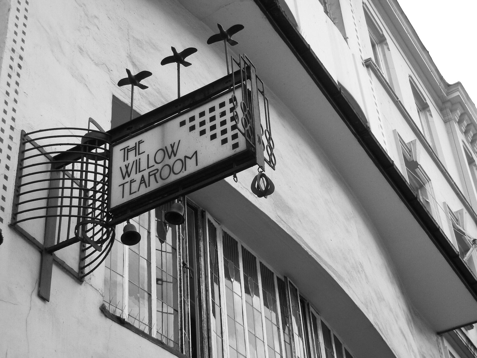 Art deco sign for "The Willow Tearoom" hanging above a vintage building, featuring decorative plant motifs in black and white.
