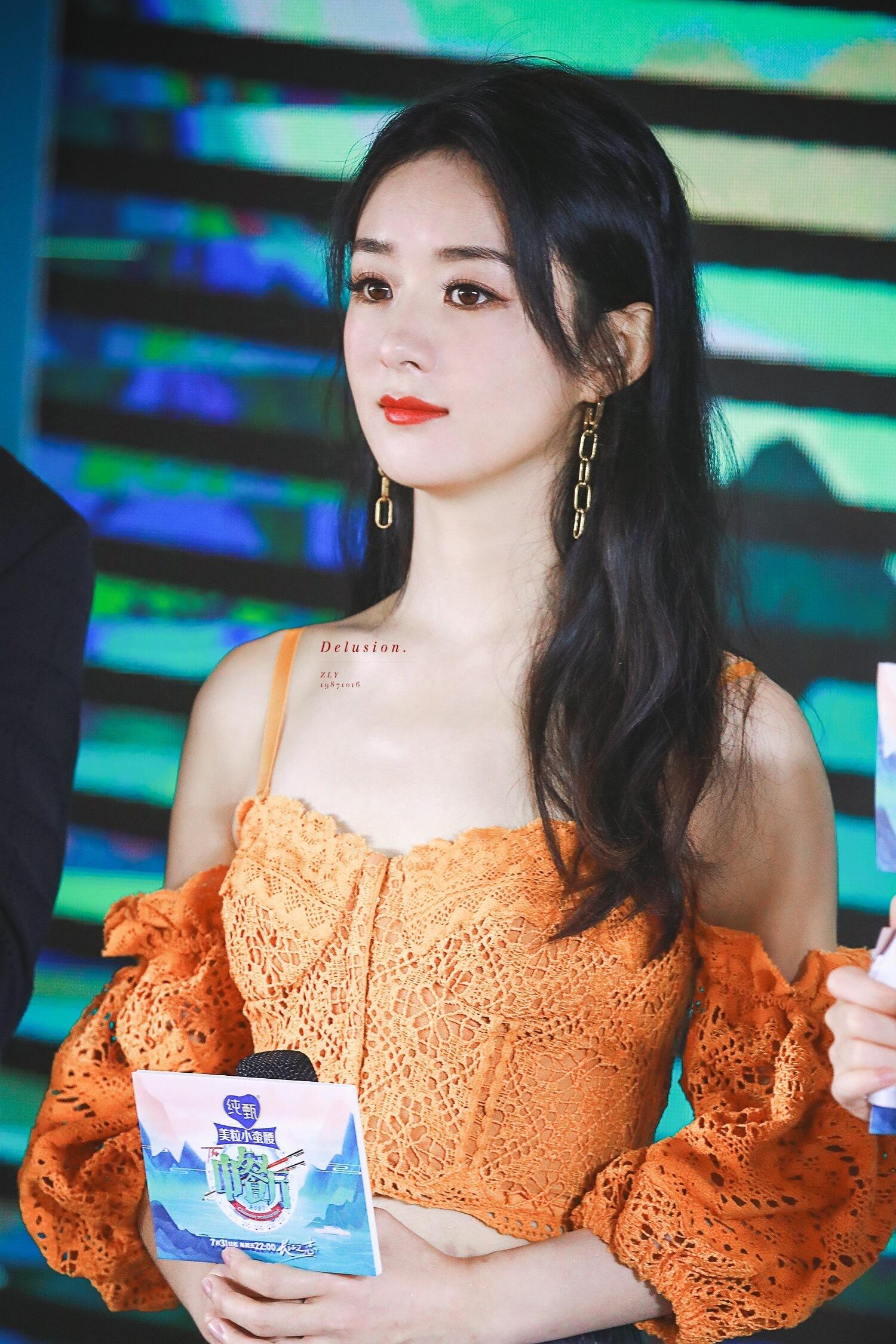 A woman with long dark hair, wearing an orange lace off-shoulder top, holds a card at an event with a colorful background.